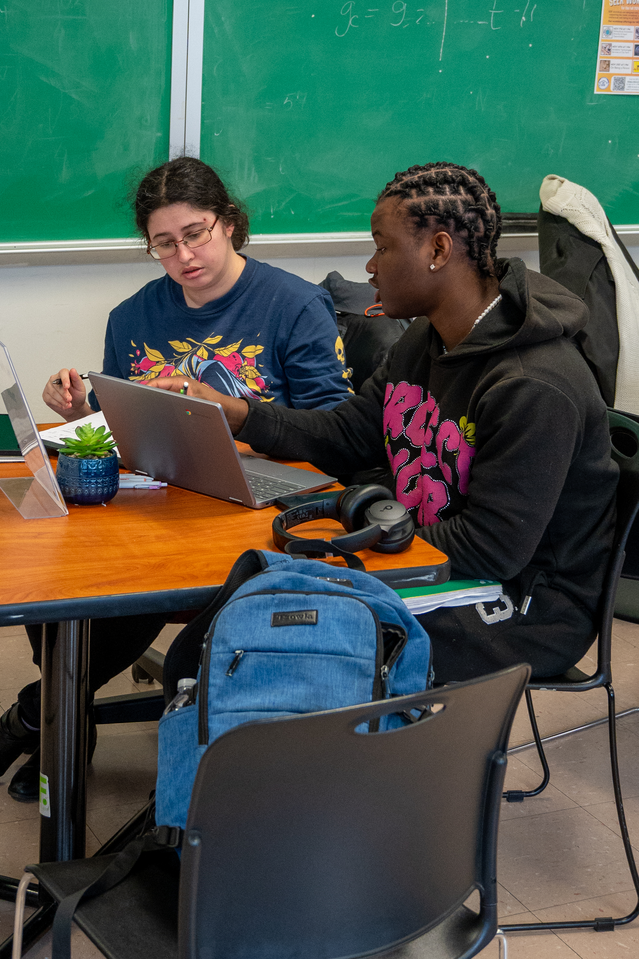 A tutor and a student sitting next to each other at a table. They are both talking while looking at the screen of a computer.