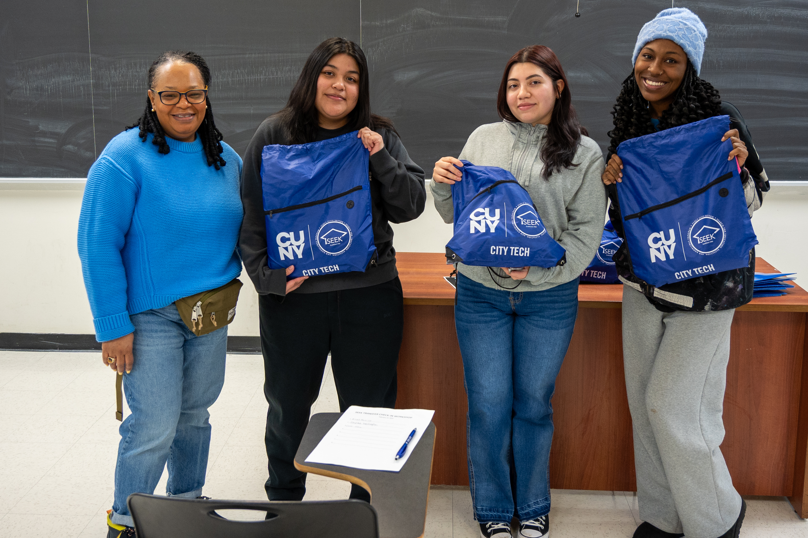 A group of three students and a professor stand in front of a classroom. The three students are holding up blue drawstring bags with the CUNY, City Tech, and CD/SEEK logos on them.