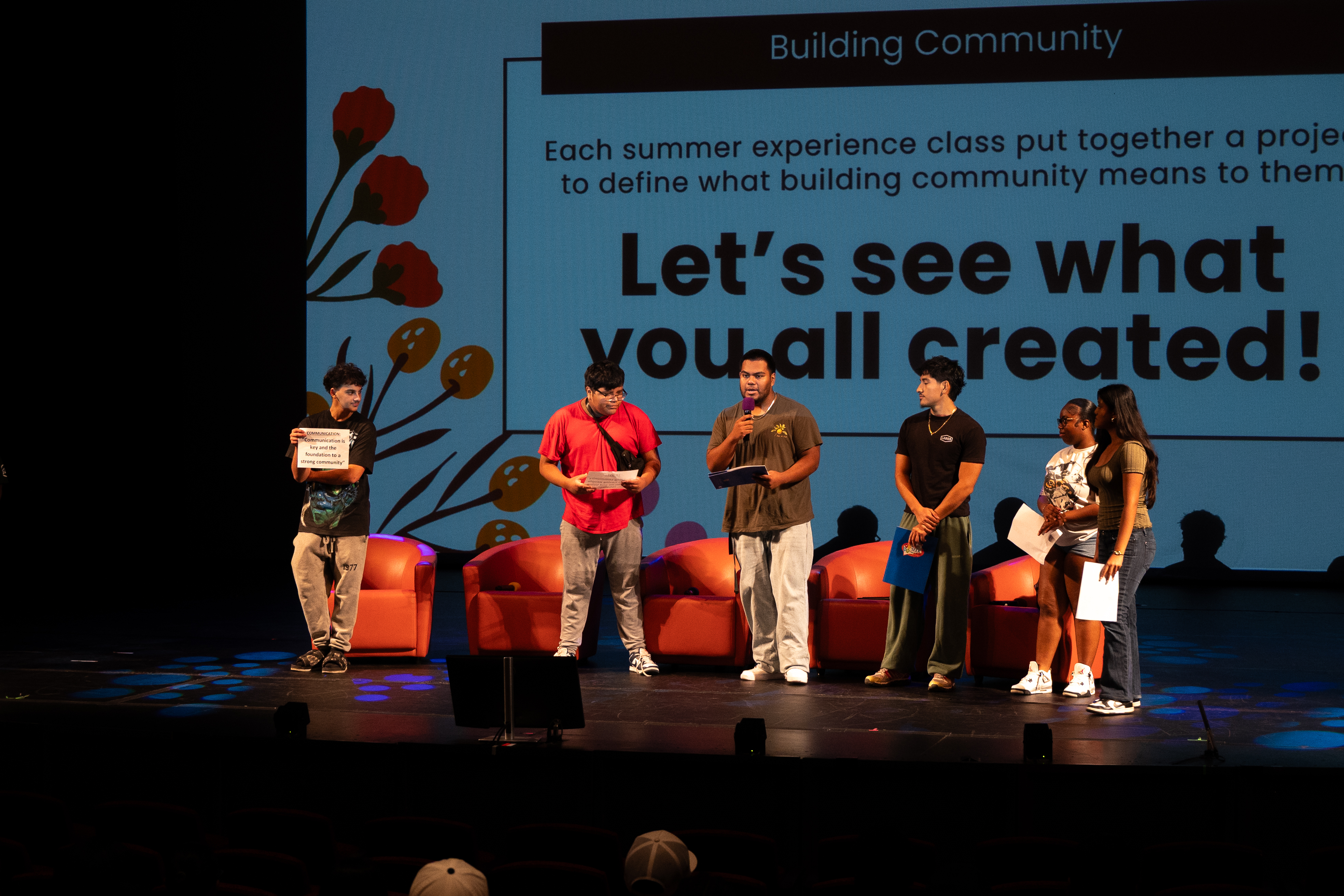 A group of students standing on a stage. One is speaking into the mic while the rest are watching. In the background, there is a PowerPoint presentation with a slide that discusses building community and showcasing student projects.