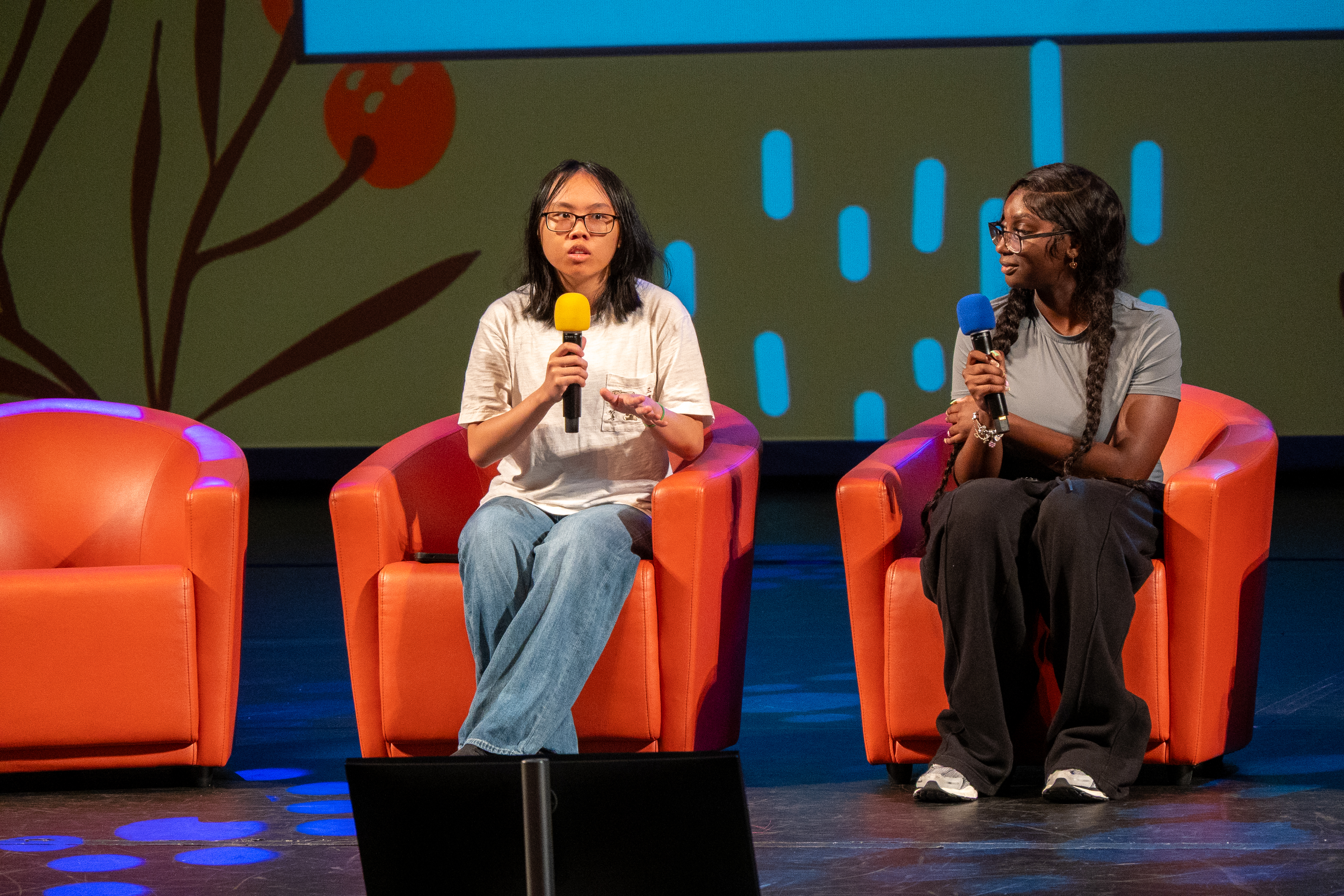 Two students sit in orange chairs on a stage. One is speaking into a microphone while the other holds a microphone and watches.